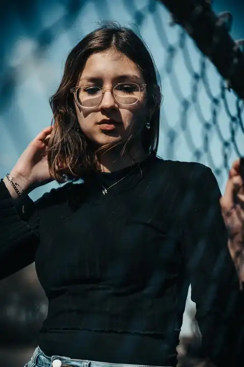 Jeune femme avec lunettes et pull noir, posant derrière un filet métallique sous un ciel bleu
